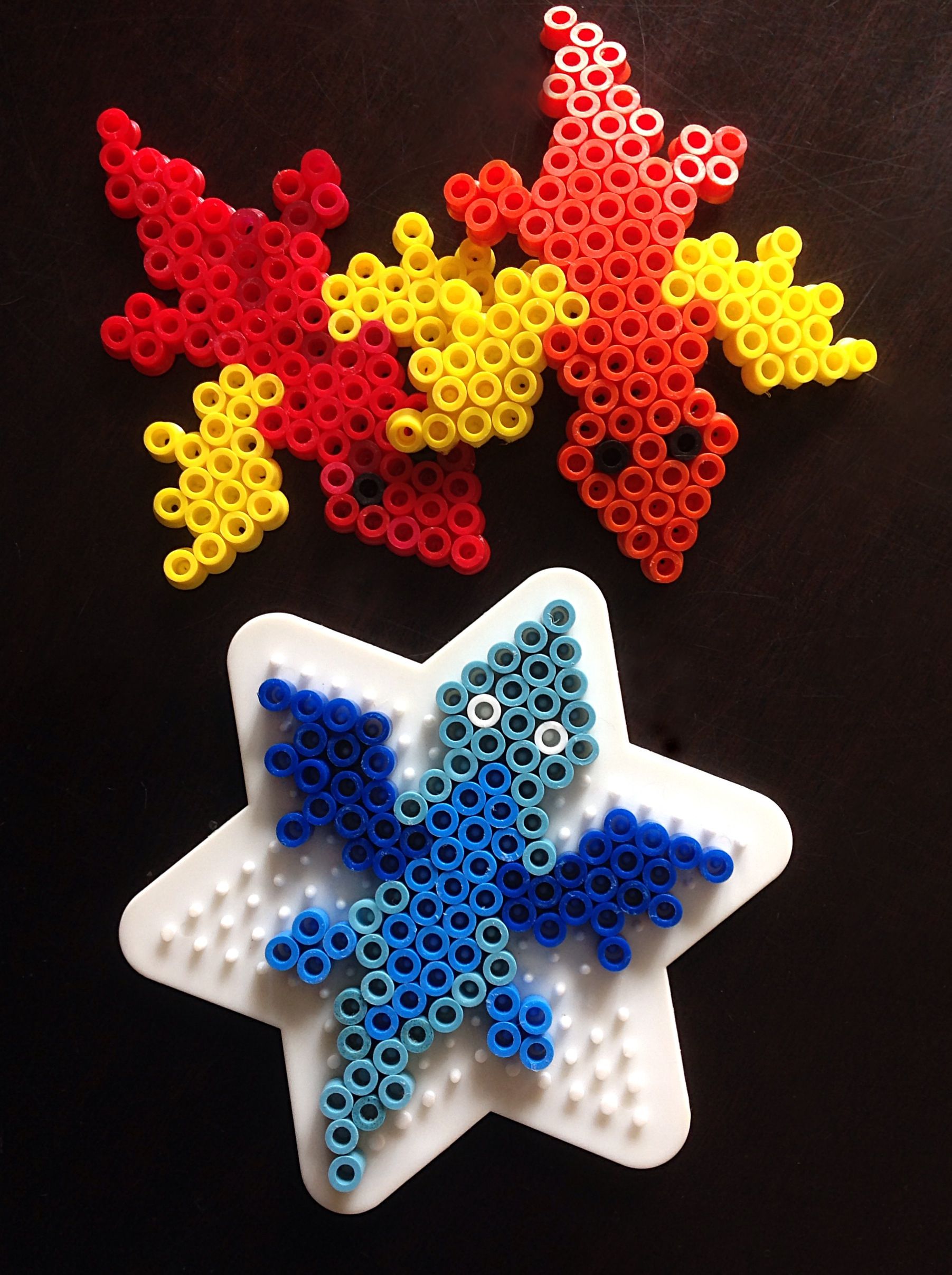 Two red and yellow dragons made from perler beads. A blue perler bead dragon still on the pegboard