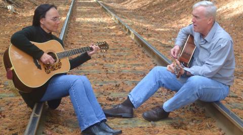 Judy and Andy Daigle sit on train tracks, each holding a guitar.