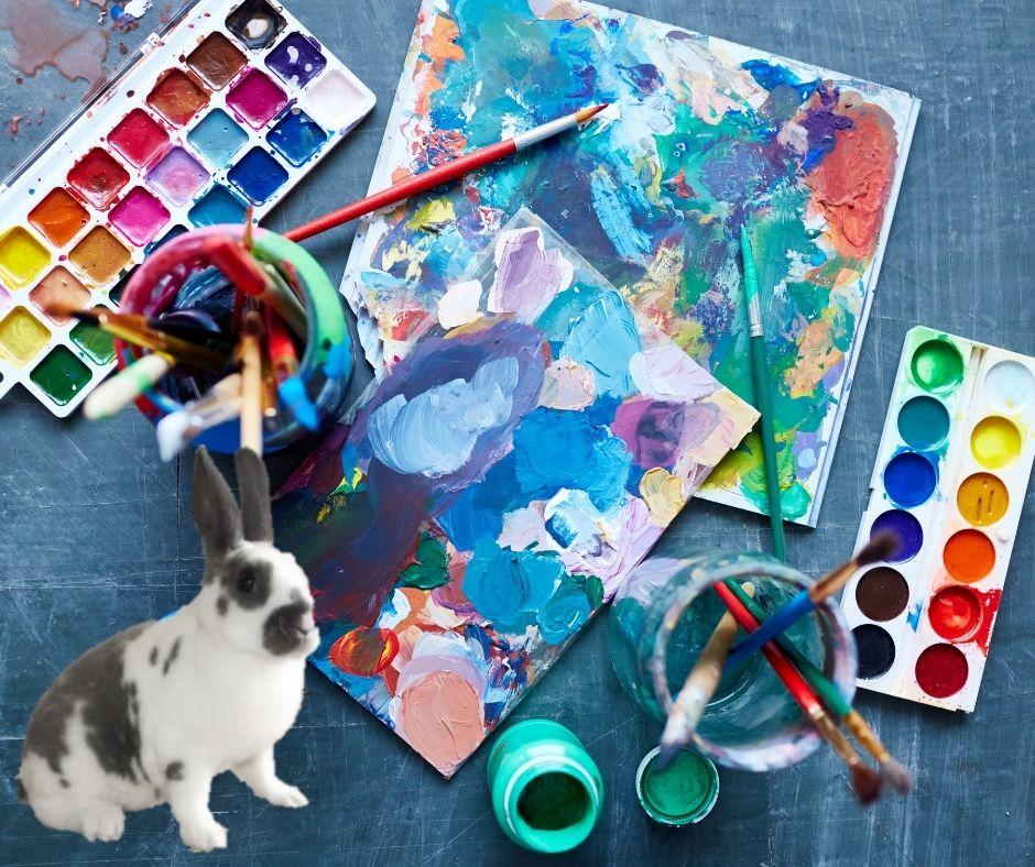 Photo of painting supplies, a canvas, water cups, and a rabbit. 